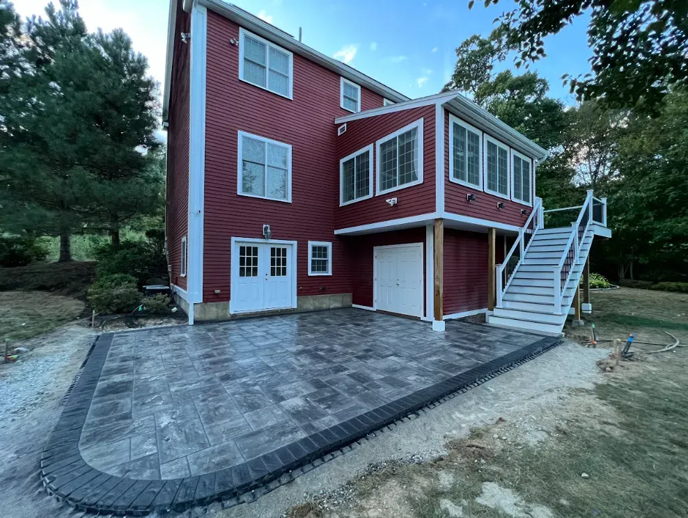 Red two-story house with paved patio, white trim, and exterior stairs in a wooded yard