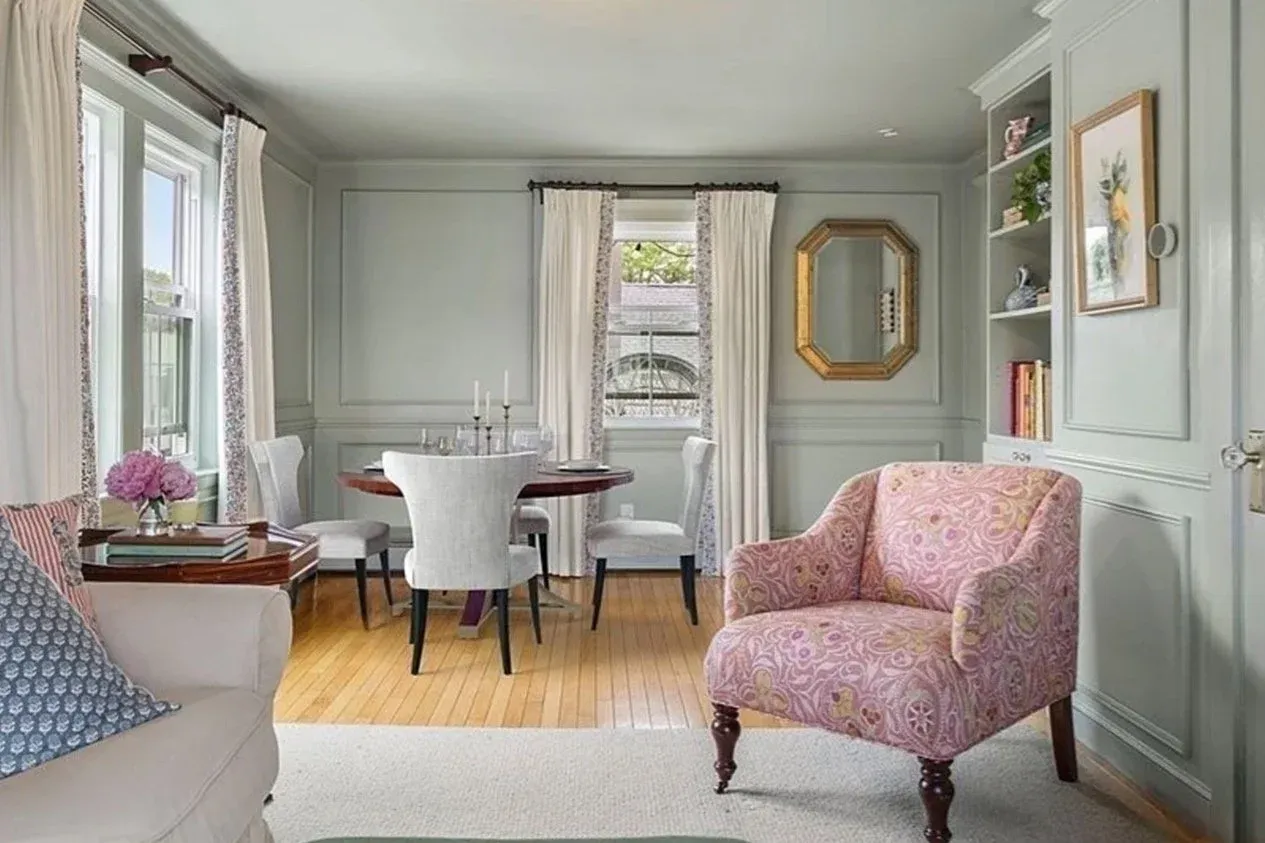 Living room with light gray walls, hardwood floor, white curtains, pink patterned armchair, sofa with patterned pillows, dining table with chairs, bookshelf, and decorative mirror and artwork.