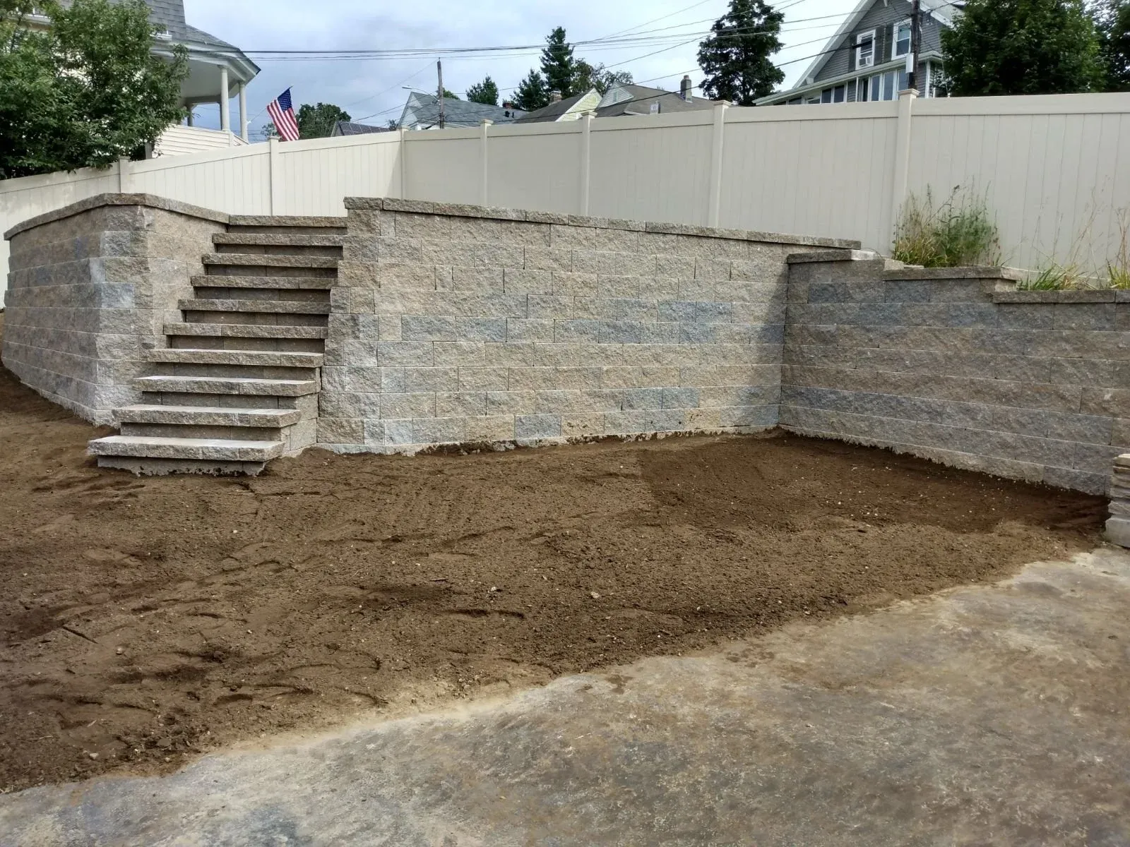 A backyard retaining wall made of gray stone blocks with stairs on the left side, under construction with dirt ground in front, fenced with white panels, houses and trees in the background.