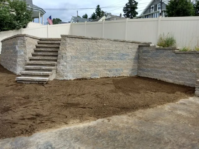A backyard retaining wall made of gray stone blocks with stairs on the left side, under construction with dirt ground in front, fenced with white panels, houses and trees in the background.