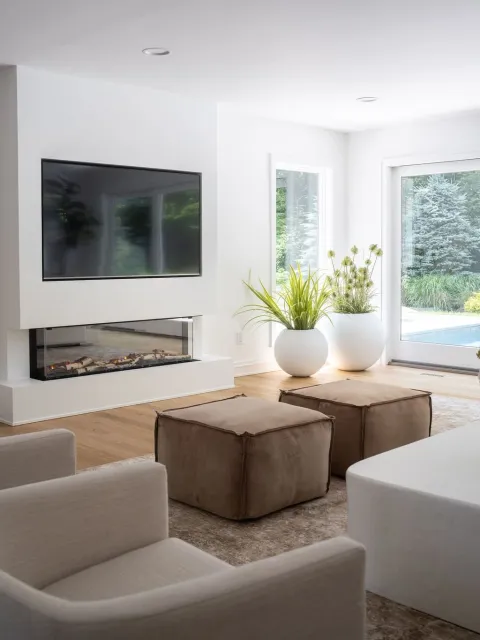 A modern living room with a wall-mounted flat-screen TV, two large white potted plants near sliding glass doors, beige ottomans, a white sofa, a fireplace below the TV, and large windows overlooking a garden with trees.