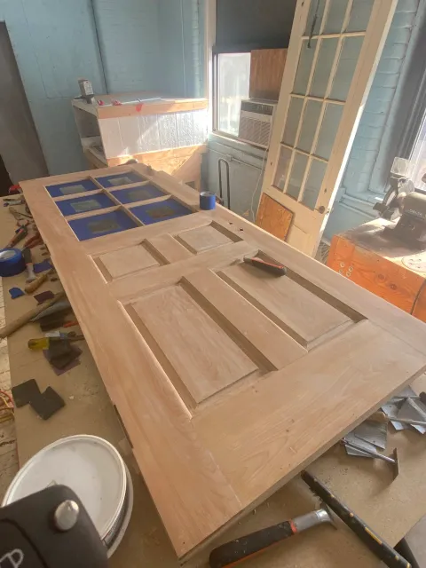 Wooden door with windowpanes being assembled on workbench in workshop, cluttered with tools and supplies, construction in progress.