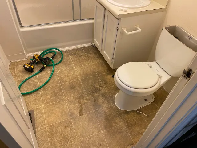 A bathroom with a shower stall, a toilet, and a small white cabinet. It appears that repair or maintenance work is being done, as there is a drill and a green hose on the floor, and water is spilled around.