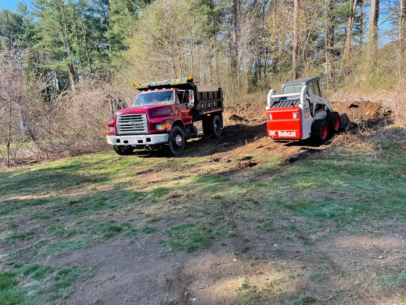 A red dump truck and a white Bobcat on a grassy hill, trees in the background.