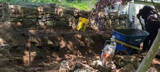 Person working in a rocky garden with a wheelbarrow and tools near a stone wall