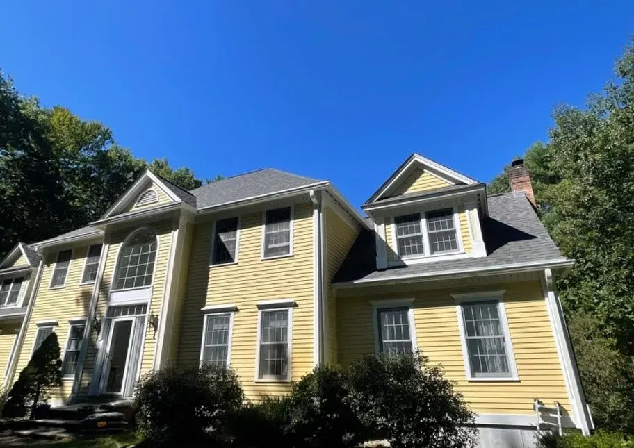 A two-story yellow suburban house with a gray shingle roof, white trim, and surrounding trees under a clear blue sky.