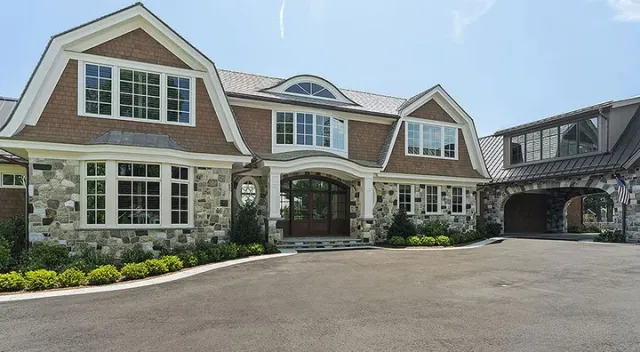 Large stone house with a curved driveway and brown roof, with clear sky visible.