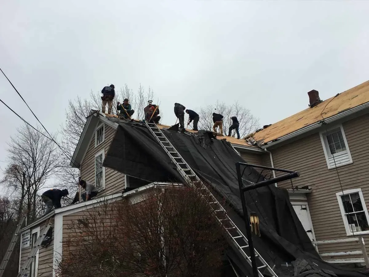 Workers repairing a steep roof, with ladders and tarps on a house in overcast weather