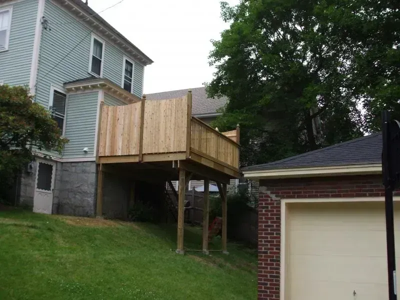 A light-green house features a newly built, elevated wooden deck with privacy walls, standing over a grassy hillside.
