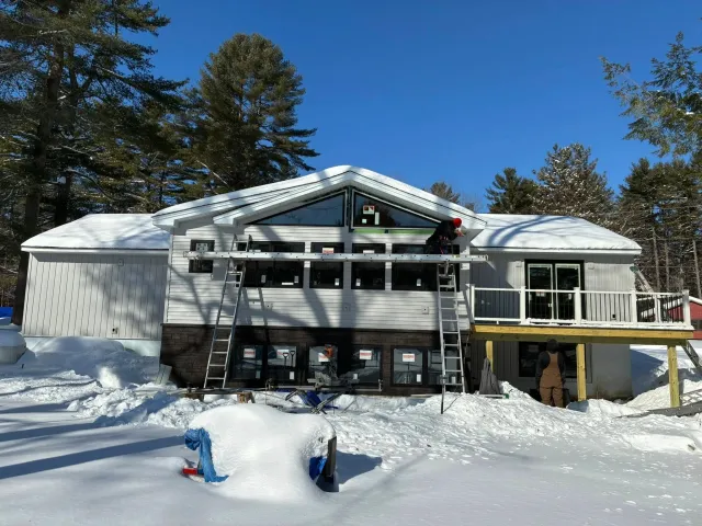 Construction workers building a house in a snowy landscape, with tall pine trees and a clear blue sky in the background.