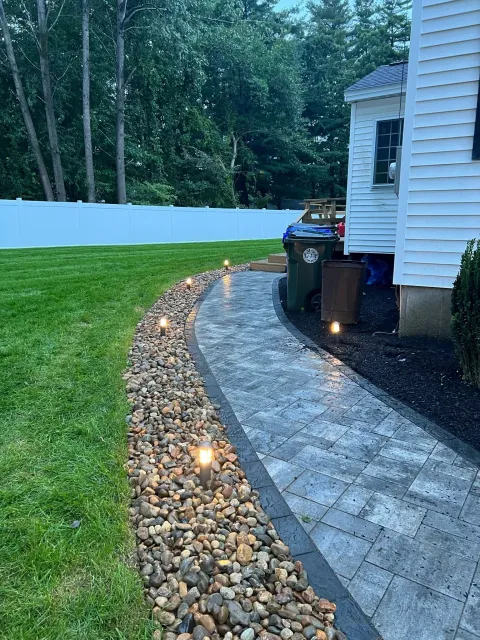 A curved stone paver walkway lit by small ground lights, bordered by a gravel bed and green lawn next to a white house.