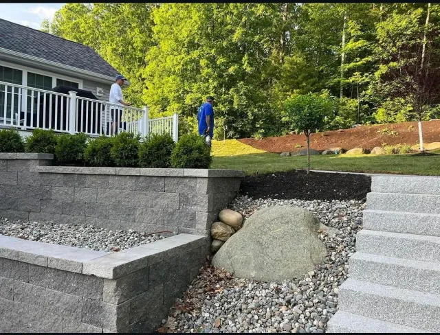 A landscaped backyard with stone retaining walls, gravel, a small tree, and two people standing on a deck overseeing the garden.