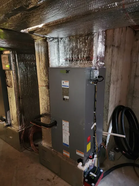 HVAC unit in a basement, ductwork is wrapped in silver insulation, next to a concrete wall.