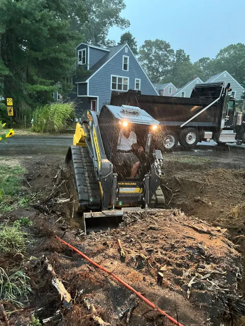 A person operates a skid steer to dig in a muddy area near a residential street, with a dump truck in the background.