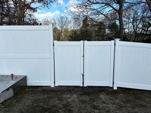 White vinyl fence with double gate in a yard with trees.