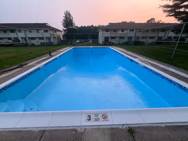 Empty swimming pool with a depth of 3 feet, surrounded by a concrete deck, fences, and residential apartments in the background during sunset.