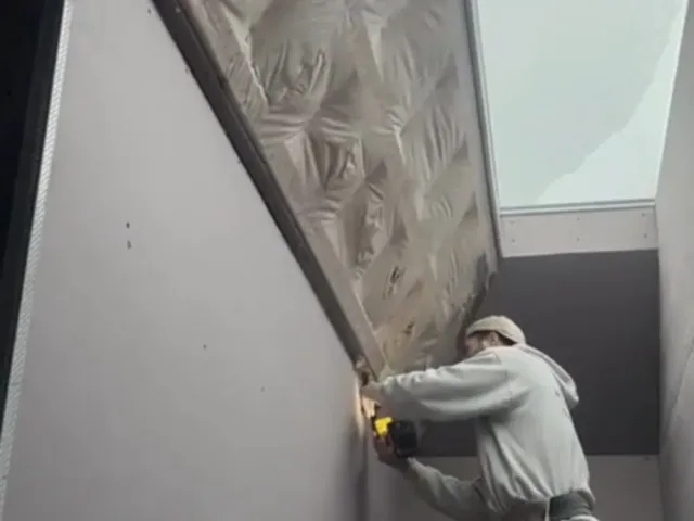 A construction worker using a screw gun on a wall during a blueboard sheetrock installation.
