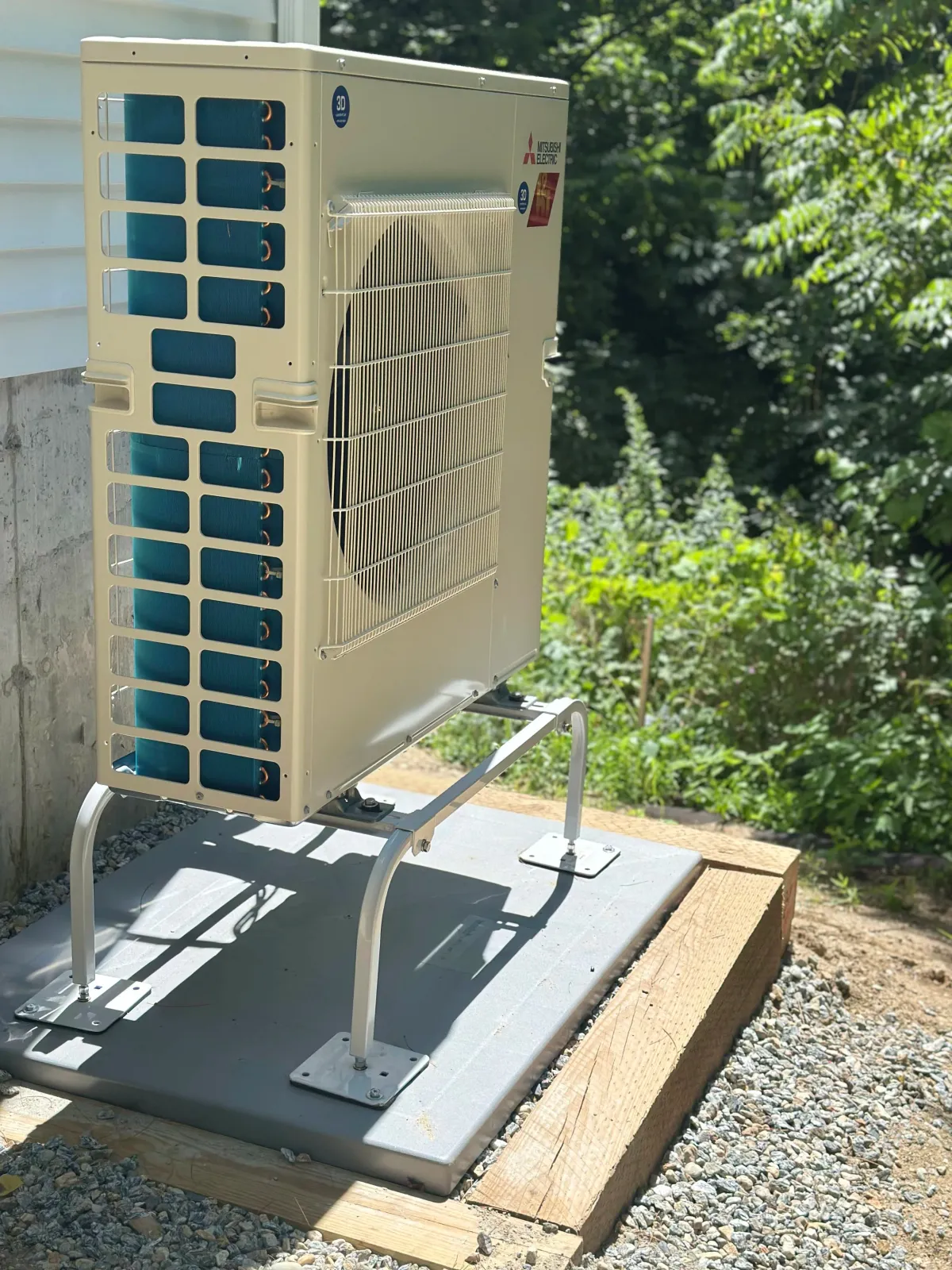 An outdoor air conditioning unit mounted on a concrete pad, with a metal stand, next to a building with white siding and surrounded by green trees and bushes.