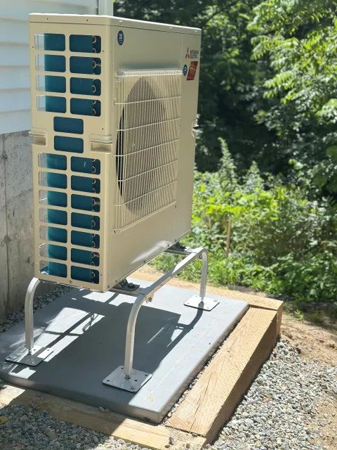 An outdoor air conditioning unit mounted on a concrete pad, with a metal stand, next to a building with white siding and surrounded by green trees and bushes.