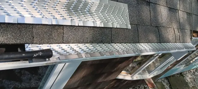 Close-up of gray roofing shingles and flashing on a sloped roof, with a patch of snow nearby