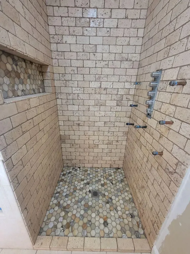 Small tiled shower stall with pebble floor, wall fixtures, and unfinished edges.
