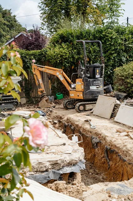A mini excavator parked beside an open footing trench in a hedged back garden