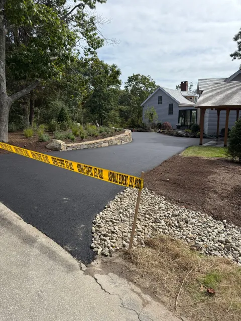 Freshly paved driveway with yellow caution tape, leading to a house with a gray exterior.