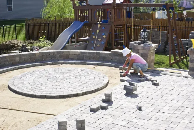 A person in a pink shirt and white hat installs stone pavers in a backyard patio near a playground set.