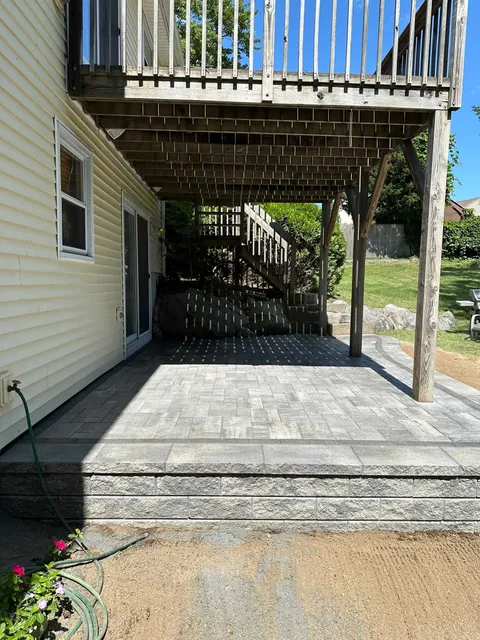 A newly installed gray paver patio beneath a wooden deck attached to the side of a light yellow house.