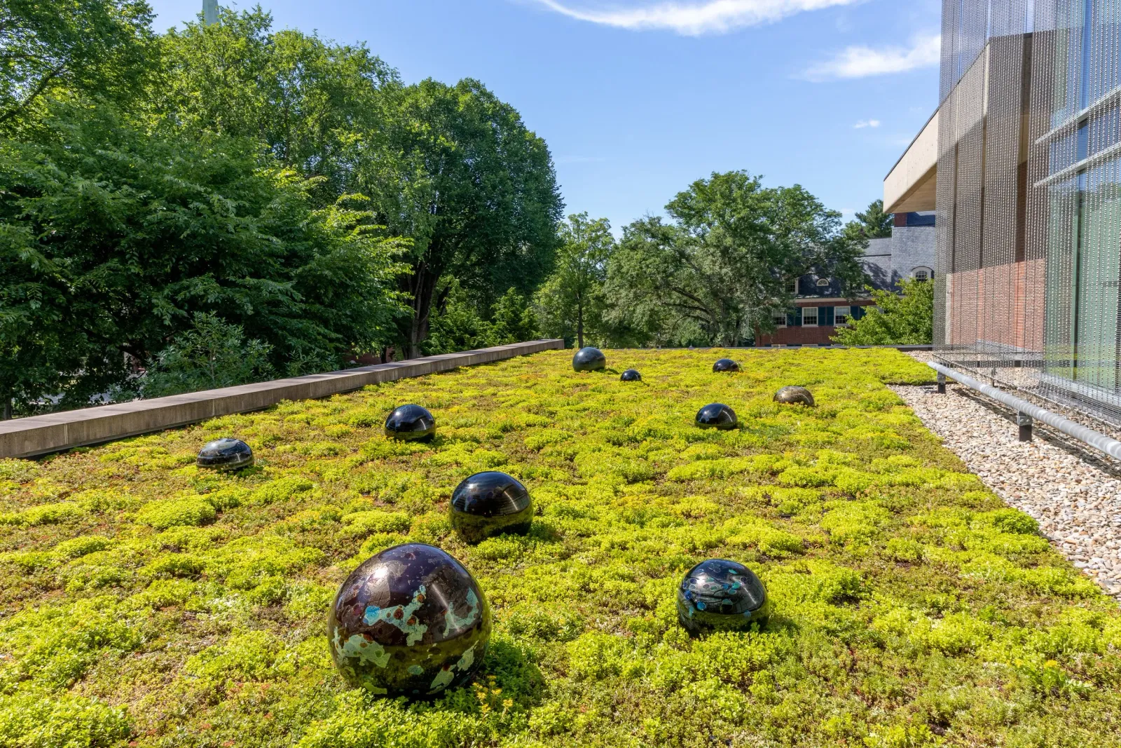 View of a green rooftop garden with decorative glass orbs, adjacent to a modern building with perforated metal panels, surrounded by trees under a blue sky with some clouds.