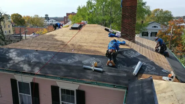 Two workers in blue shirts install black underlayment on the wooden plywood roof of a pink house.