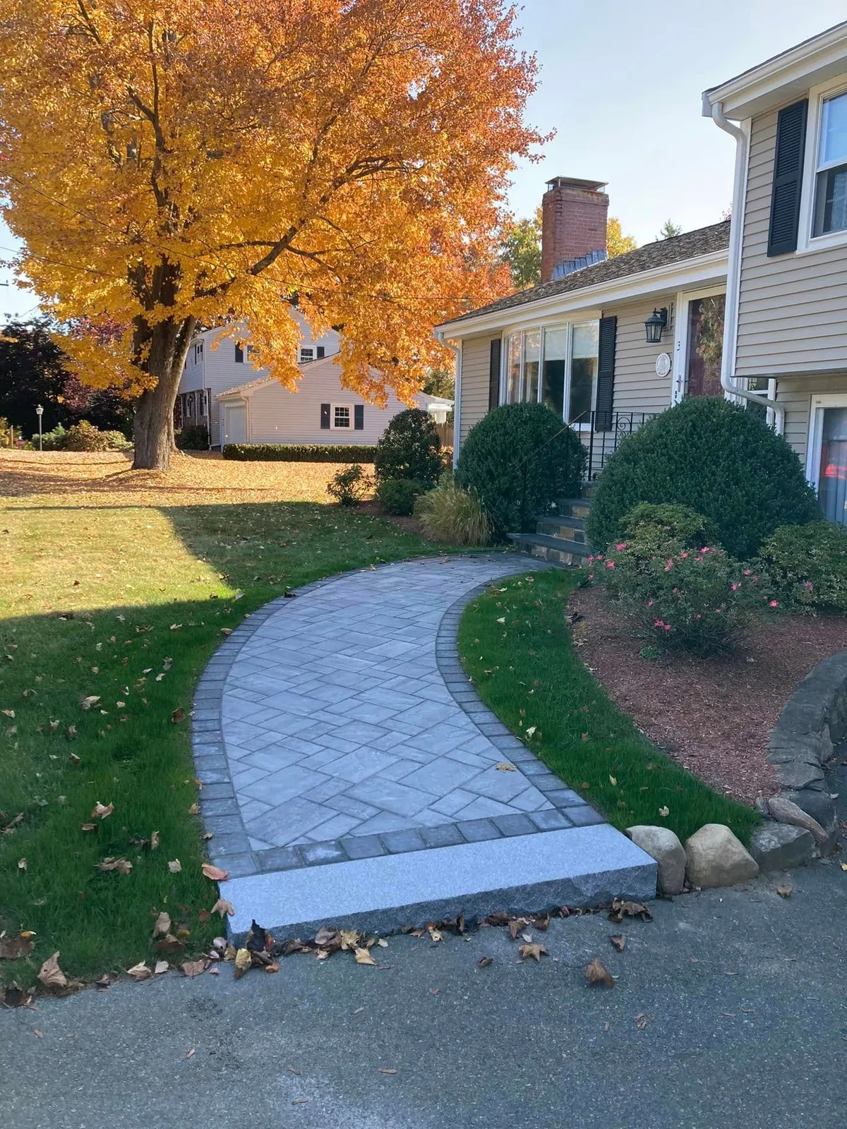 A stone walkway curves from a driveway to a house with autumn foliage.
