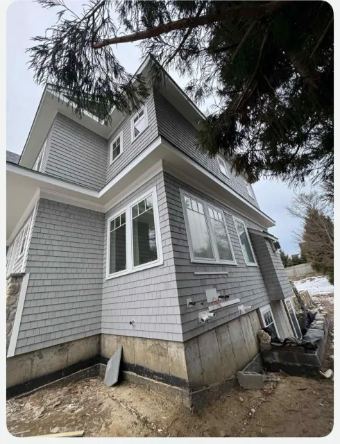 Gray house exterior with damaged foundation and leaning side wall under overhanging tree branches