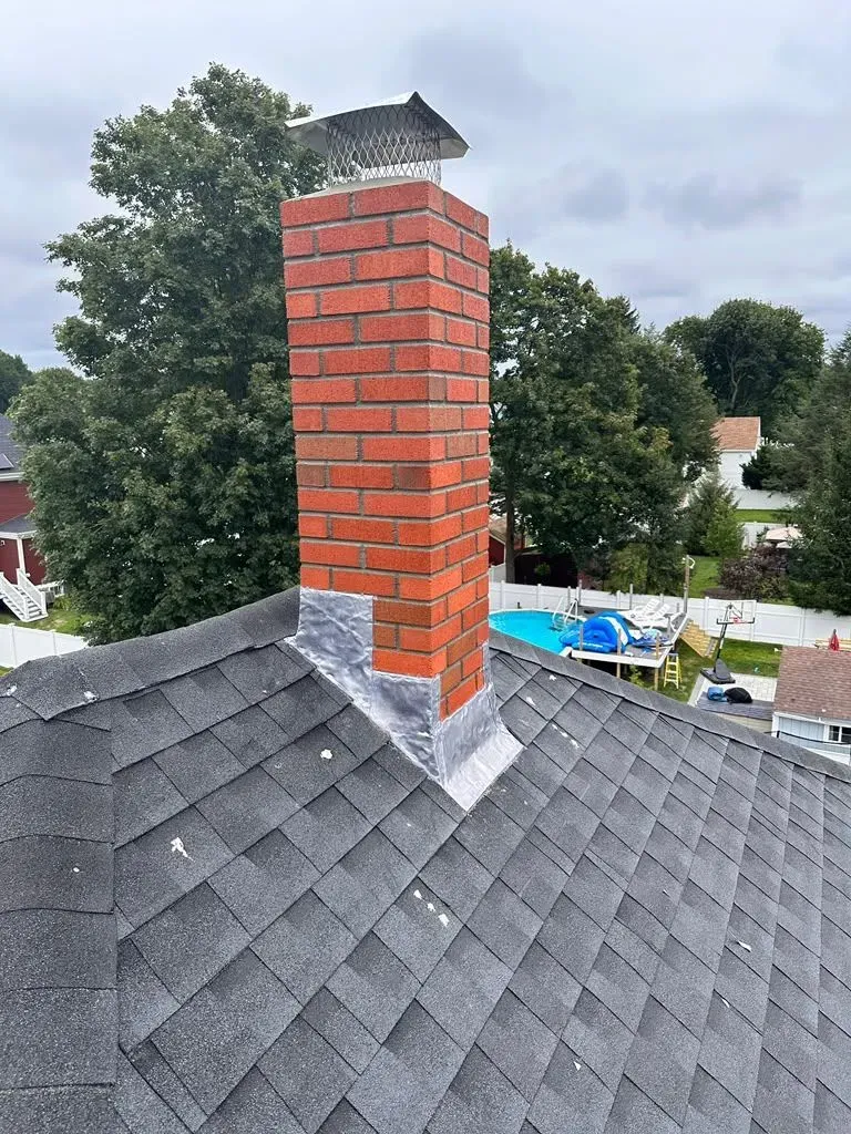Red brick chimney on a gray shingled roof with nearby trees and cloudy sky
