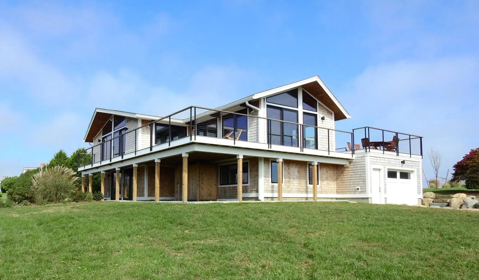 A modern two-story house with a large balcony, glass railings, and a mix of wood and white exterior, set in a green yard with trees and rocks, under a blue sky.