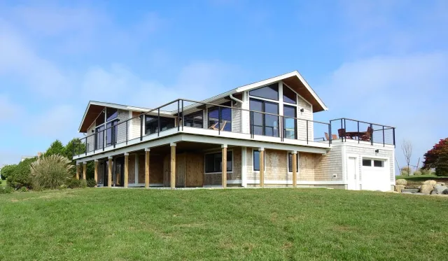 A modern two-story house with a large balcony, glass railings, and a mix of wood and white exterior, set in a green yard with trees and rocks, under a blue sky.