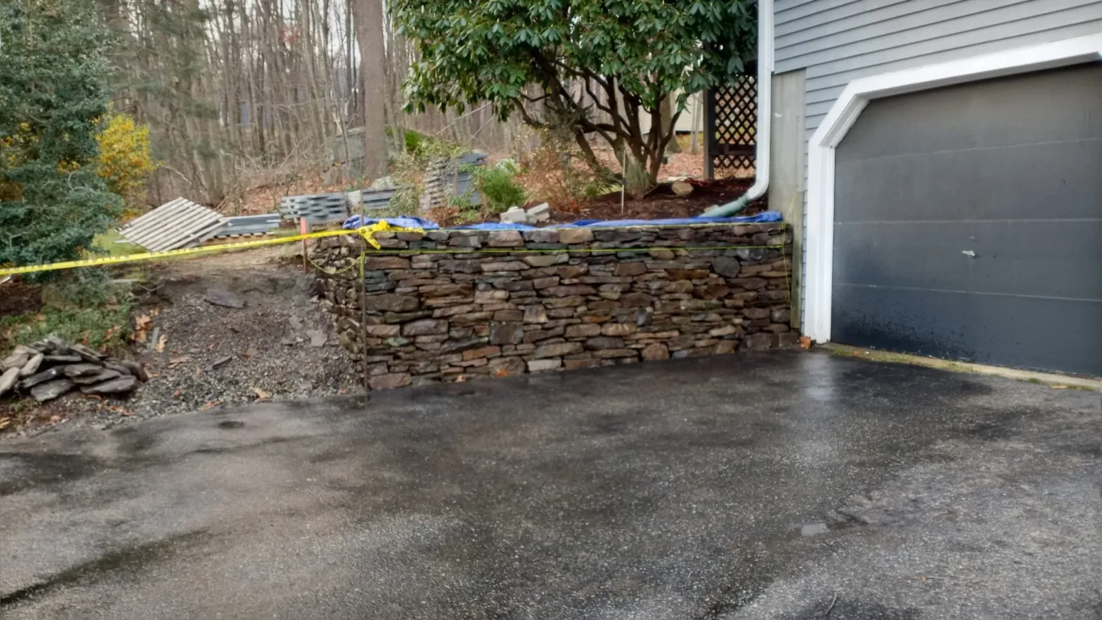 A newly built stone retaining wall beside a driveway, with construction materials and yellow caution tape nearby, in a residential area with trees in the background.