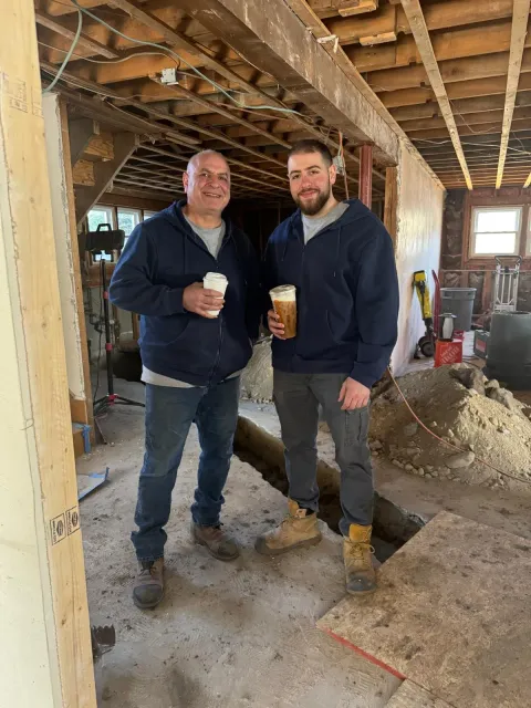 Two men standing inside a construction site holding drinks and smiling at the camera. The man on the left is older and wearing a navy hoodie and jeans, while the man on the right is younger with a beard, wearing a navy hoodie, cargo pants, and work boots. There are construction materials and tools around, and the ceiling is unfinished.