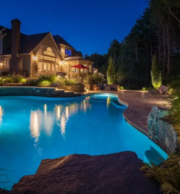 Night view of a lit, curved swimming pool in a luxury backyard with a illuminated house and outdoor seating area.