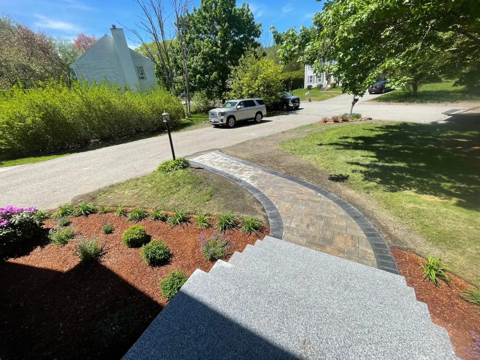 Curved stone path through a sunny garden with red mulch, shrubs, trees, and a parked white SUV nearby.