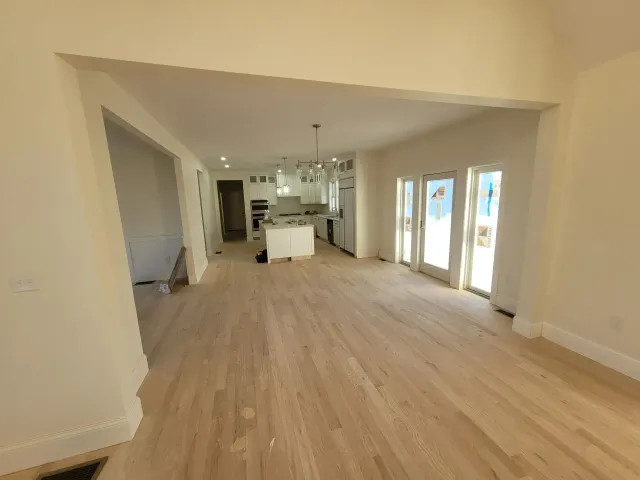Spacious interior with light wood floors, leading to a kitchen with white cabinets. Sunlight streams in from glass doors.
