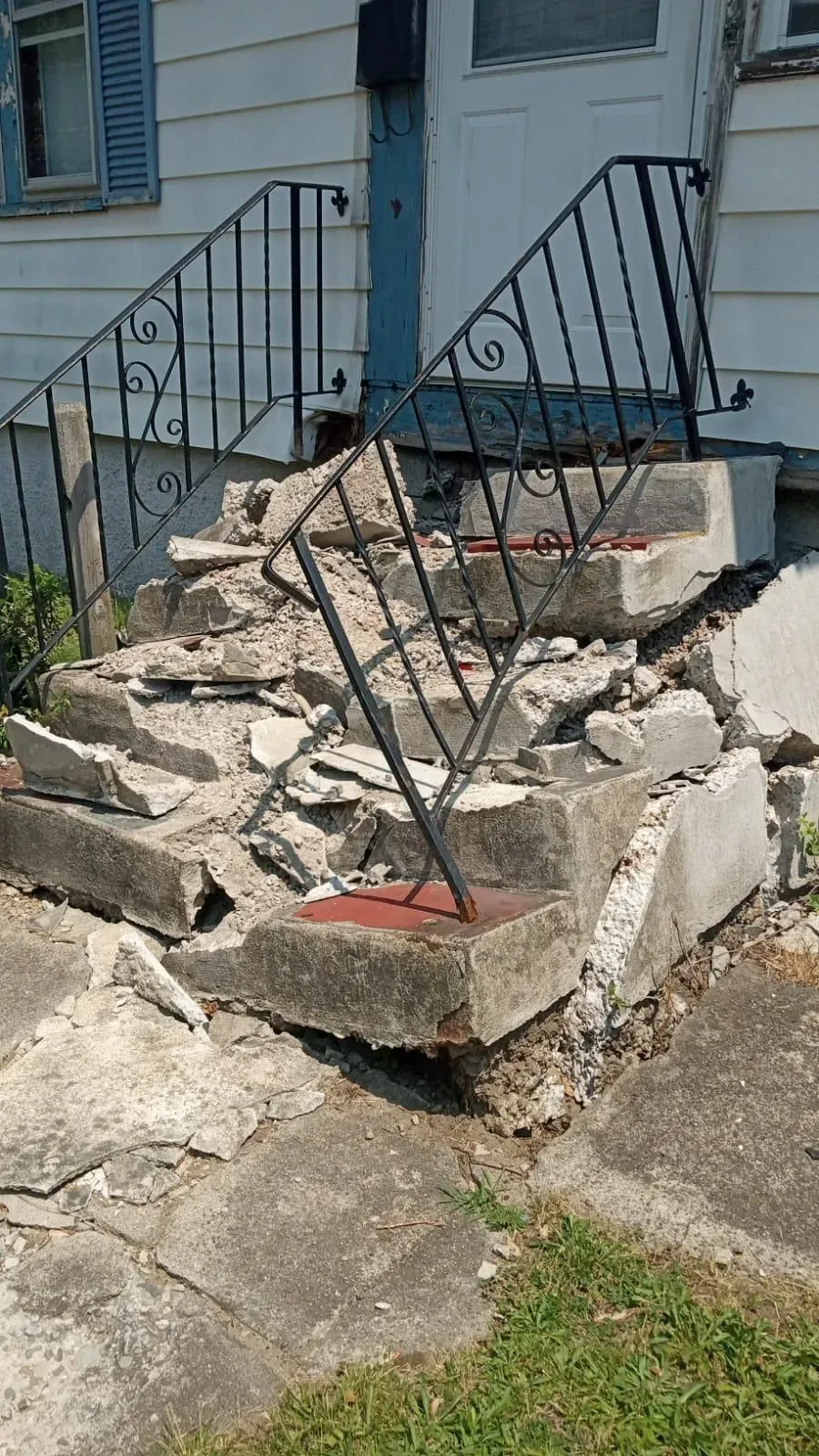 Broken concrete porch steps with a bent black metal railing outside a house