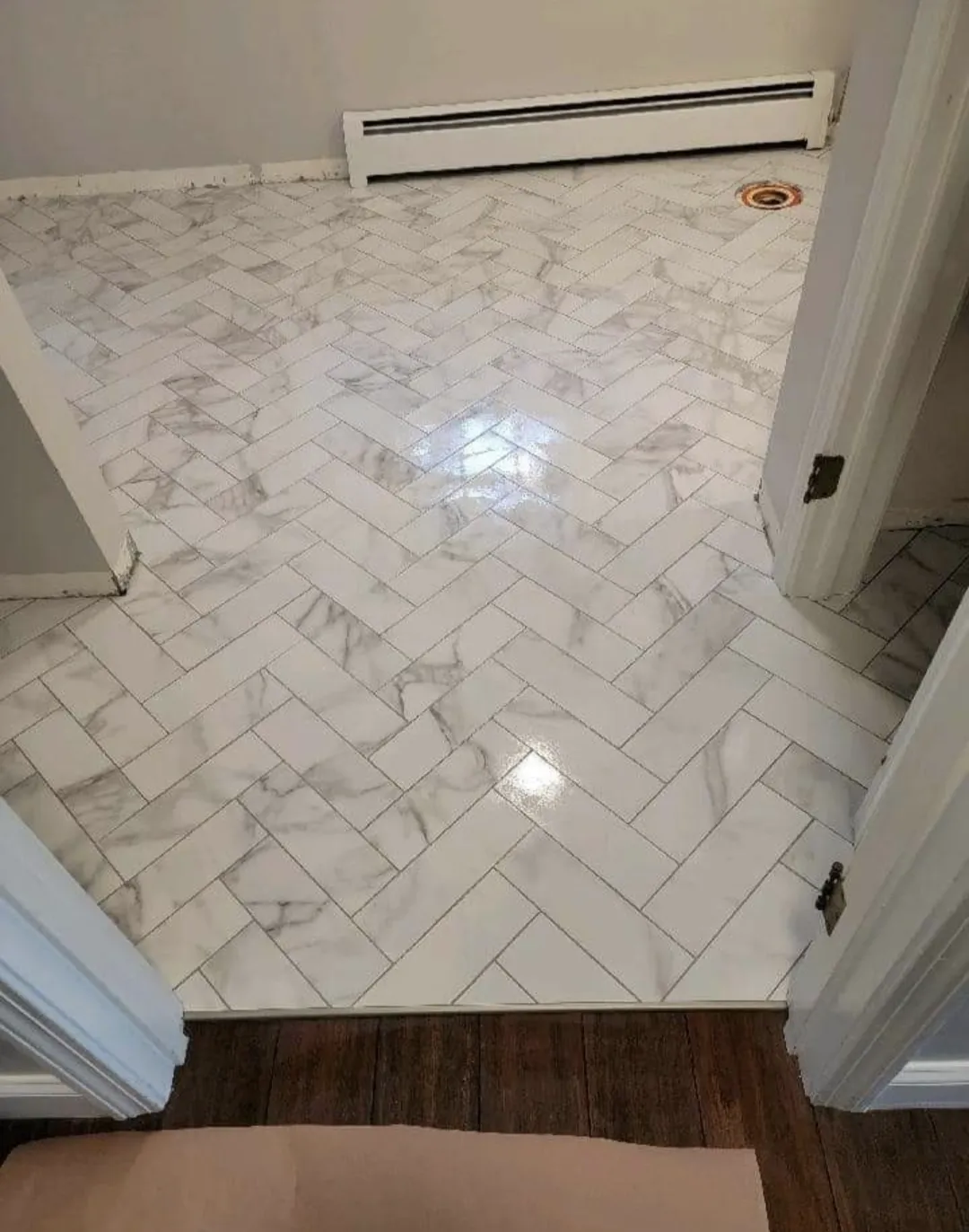Newly tiled white marble herringbone pattern floor in a room, with baseboard heater and vent in the background.