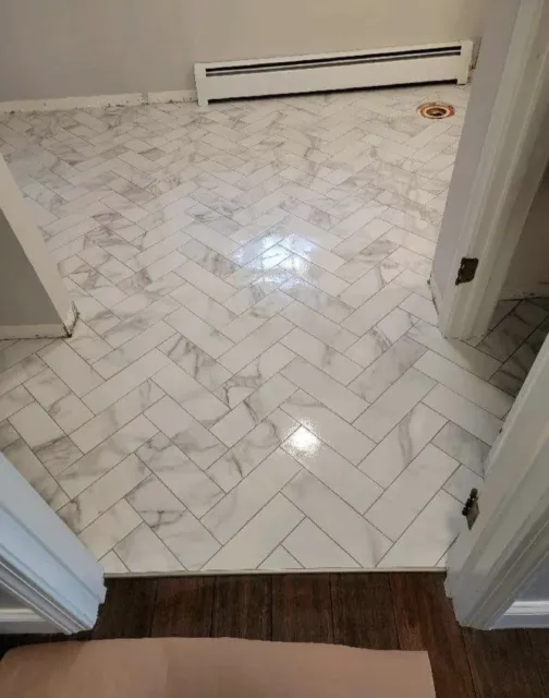 Newly tiled white marble herringbone pattern floor in a room, with baseboard heater and vent in the background.