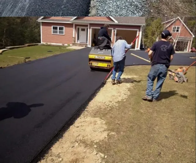 Construction workers pave a new asphalt driveway in front of a house, using a small yellow roller and hand tools.