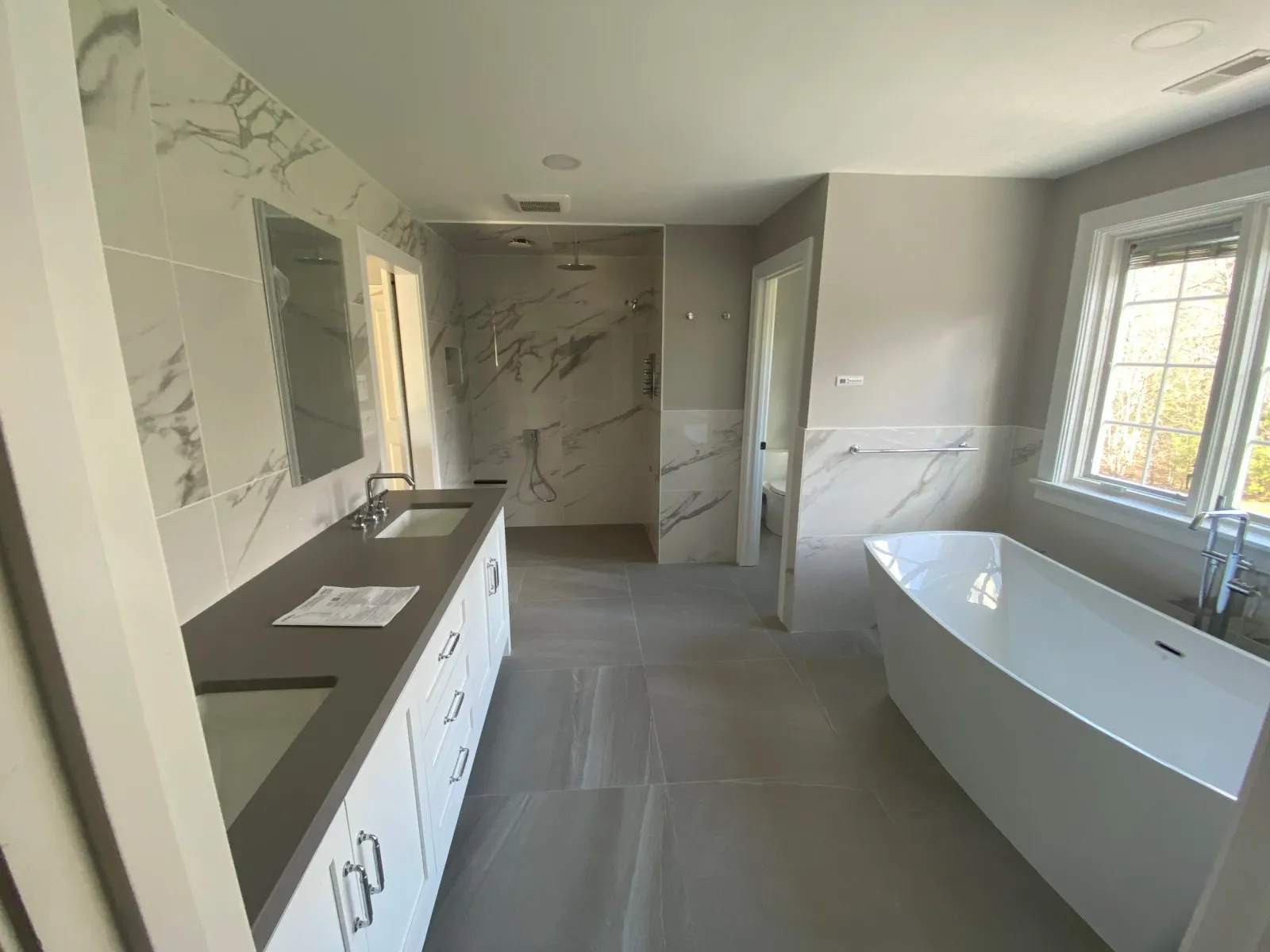 Bathroom featuring a modern bathtub by a large window, a double vanity with two sinks, and a walk-in shower in the background with marble tiles.
