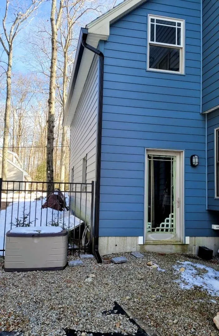 Back side of a blue house with a sliding glass door and a window, black downspout, and a small fenced area with snow, a shed, and gravel ground.