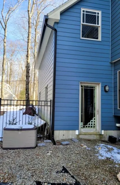 Back side of a blue house with a sliding glass door and a window, black downspout, and a small fenced area with snow, a shed, and gravel ground.