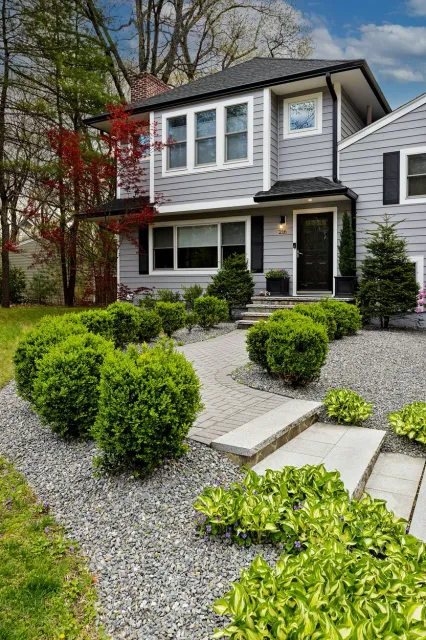 Front yard of a gray two-story house with a black door, surrounded by neatly trimmed bushes and small trees, with a stone pathway leading to the entrance.