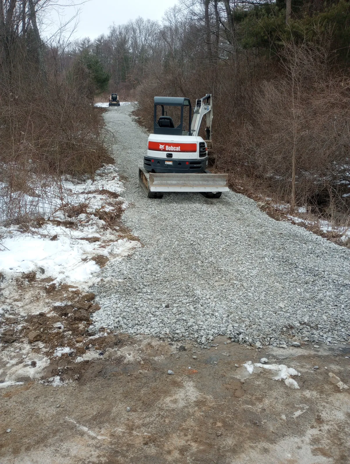 A small Bobcat excavator parked on a gravel trail in a wooded area, with snow and bare trees on both sides.